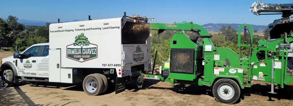 A white commercial truck with "Familia Chavez Tree Service and Tree Trimming" logo parked next to a green industrial wood chipper in a rural setting.