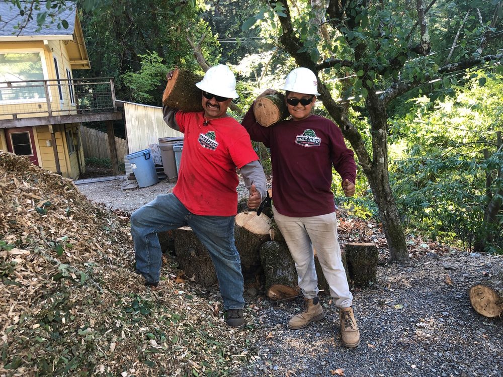 Two workers in hard hats posing after emergency tree service with a fallen tree and wood chips.